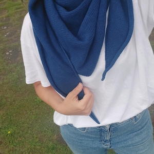 May include: A navy blue knit scarf draped over a white t-shirt. The scarf is triangular and appears to be made of a soft, textured material. The person is wearing blue jeans. The background is a mix of green grass and a paved surface.