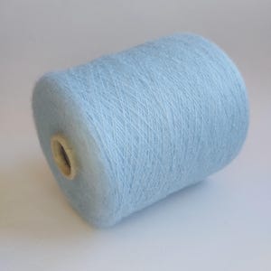 May include: A cylindrical spool of light blue yarn with a soft, fuzzy texture. The yarn is tightly wound around a central cardboard core, displaying a consistent pale blue color throughout the entire length of the thread on the spool.