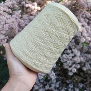 May include: A large spool of pale green yarn held in a hand. The yarn is tightly wound, showing a crisscross pattern. The background features soft pink and green foliage, suggesting an outdoor setting. Ideal for knitting and crochet projects.