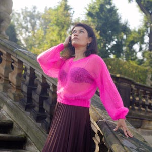 May include: A woman wearing a bright pink, sheer, knitted sweater with a brown pleated skirt. She is standing on a stone staircase with a stone railing.