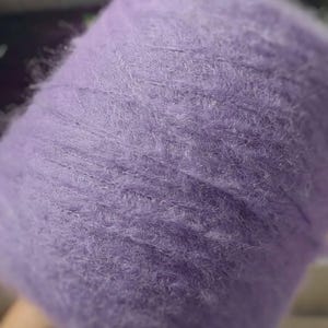 May include: A close-up of a spool of lavender-colored yarn. The yarn appears soft and fluffy, with a slightly textured surface. The color is a muted purple, and the yarn is tightly wound around the spool. This yarn is suitable for knitting and crocheting projects.