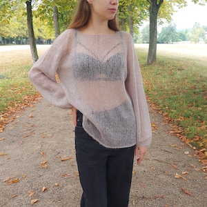 Könnte beinhalten: Ein durchsichtiger, hellrosa Pullover mit lockerer Passform. Der Pullover ist aus zartem Strick gefertigt und hat einen leicht oversized Look. Das Model trägt den Pullover zu schwarzen Jeans und weißen Sneakers.