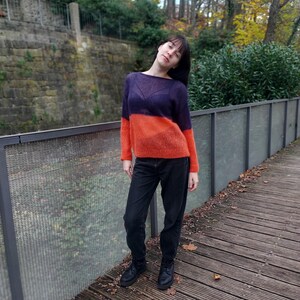 Puede incluir: Una mujer con un suéter de dos tonos naranja y morado con jeans negros y botas negras. Está de pie en una pasarela de madera con una valla de metal detrás de ella.