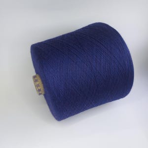 Puede incluir: Una gran bobina de hilo azul marino. El hilo está bien enrollado y tiene una textura suave. Una pequeña etiqueta beige con estrellas doradas está adherida al lado de la bobina. El hilo es adecuado para tejer, hacer ganchillo y otras manualidades.