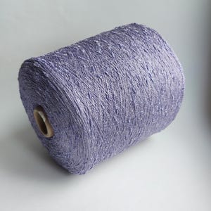 May include: A large cylindrical spool of light purple yarn with a textured, slightly metallic appearance. The yarn is wound tightly around a central cardboard core, displaying a consistent lavender hue and a nubby, uneven surface texture throughout.