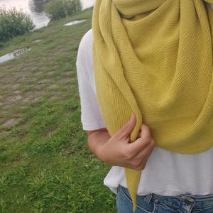 May include: A mustard yellow knitted scarf with a triangular shape. The scarf is draped around a person's neck and shoulders.