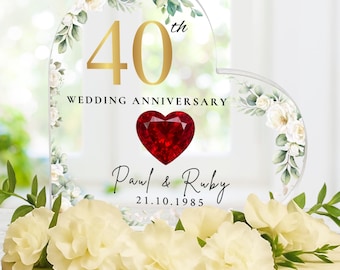 May include: A cake topper for a 40th wedding anniversary. The clear acrylic heart has gold lettering, a red heart, and the names "Paul & Ruby" with the date 21.10.1985. The cake is decorated with white flowers and greenery.