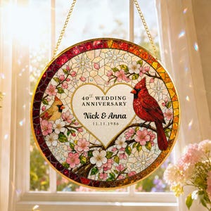 May include: A circular stained glass suncatcher featuring a red cardinal and a yellow bird perched on branches with pink blossoms. A central heart contains the text 40th WEDDING ANNIVERSARY, Nick & Anna, and 11.11.1986. It hangs from a gold chain.