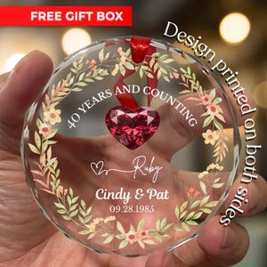 May include: A clear, faceted glass ornament with a red heart-shaped gem in the center. The ornament is decorated with floral designs and the text "40 YEARS AND COUNTING", "Ruby", "Cindy & Pat", and "09.28.1985". A red ribbon is attached for hanging.