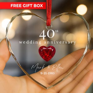 May include: A clear, heart-shaped crystal ornament with the text "40th wedding anniversary." A red heart-shaped gem is in the center. The names "Mark & Ellen" and the date "9-21-1985" are also visible. A red ribbon is attached.