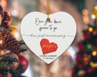 May include: White heart-shaped ceramic ornament with the text "One year down, forever to go. Our first anniversary paper" and a red origami paper heart. The ornament also includes the names "GEORGE & MERRY" and the date "17.09.2023".