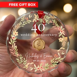 Puede incluir: Un adorno de cristal transparente y facetado con diseños florales y de hojas. El adorno tiene el texto "40 aniversario de bodas", junto con nombres y una fecha. También son visibles una cinta roja y un sello de cera dorado con una rosa.