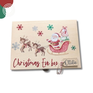 May include: A rectangular wooden box featuring a printed illustration of Santa Claus in a red sleigh pulled by two reindeer. The box is decorated with red, green, and silver snowflakes and the text "Christmas Eve box" with a wooden tag labelled "Ottilie".