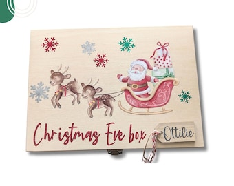May include: A rectangular wooden box featuring a printed illustration of Santa Claus in a red sleigh pulled by two reindeer. The box is decorated with red, green, and silver snowflakes and the text "Christmas Eve box" with a wooden tag labeled "Ottilie".