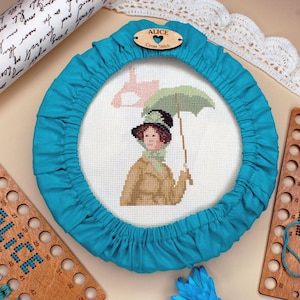 Może przedstawiać: Obręcz do haftu krzyżykowego z turkusową osłoną z tkaniny, z ukończonym wzorem haftu krzyżykowego kobiety z parasolem. Widoczne są również drewniane szpulki i zwój z tekstem. Projekt oznaczony jest jako "ALICE".