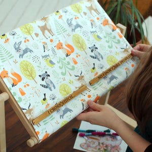 May include: A cross-stitch frame made of wood, displaying fabric with a woodland animal design. The fabric shows deer, foxes, owls, and raccoons. The wooden bars have the text "I CROSS STITCH... SO I DON'T KILL PEOPLE".