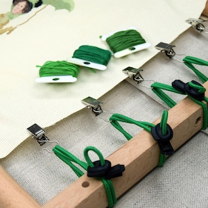 May include: A wooden embroidery hoop with a canvas stretched across it, secured by metal clips and green elastic bands. Three spools of green embroidery floss are placed on the canvas, next to a partially stitched design.