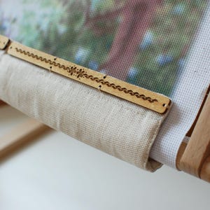 May include: A wooden embroidery frame holding canvas and fabric. The wooden frame has decorative cutouts. The canvas displays a detailed, colourful image. The fabric is a light beige colour.