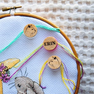 Peut inclure: Un cercle à broder avec un motif de lapin et des détails floraux. Des marqueurs de fil en bois avec des silhouettes de chat et le nom "ERIN" sont attachés au tissu avec des fils colorés. Le cercle est en bois.