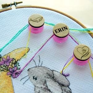 May include: Cross-stitch embroidery hoop featuring a bunny design and colourful threads. Three wooden bobbins with pink centres are visible, one with the name "ERIN" and the others with whale and heart designs. The embroidery is in progress.