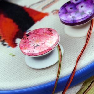 May include: Close-up of two decorative needle minders with floral designs. One is pink with white flowers, the other is purple with a floral pattern. They are attached to a white fabric stretched on a blue embroidery hoop, with colorful threads.