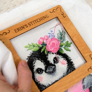 Könnte beinhalten: Kreuzstich-Stickerei eines Pinguins mit Blumenkrone, eingerahmt in einem hölzernen Stickrahmen. Der Rahmen trägt die Aufschrift "ERIN'S STITCHING". Der Pinguin hat schwarz-weiße Federn, rosa Wangen und einen rosa-grauen Körper.