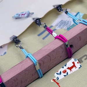 Könnte beinhalten: Ein Holzkleiderbügel mit Metallklammern und rosa und blauen Gummibändern. Ein Anhänger mit einer Fuchsgrafik hängt am Bügel. Der Bügel hält einen Stoff mit Muster.