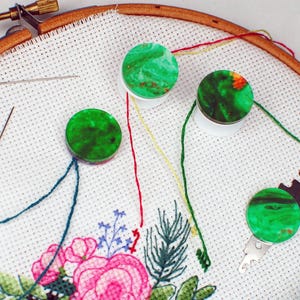 May include: An embroidery hoop with a floral design in pink, green, and blue. Green thread spools and needles are placed on the white fabric. A metal needle threader is also visible. The hoop is made of wood.