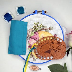 May include: A cross-stitch embroidery kit with a floral design, a wooden fox-shaped thread organizer, and blue embroidery floss. A partially stitched design is in a blue hoop. A teal fabric and a pink and white rose are also present.