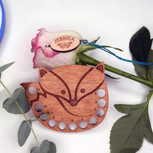 May include: A wooden fox-shaped embroidery floss organizer with eleven holes, a rose, and a small wooden tag with the name "Jessica" and a butterfly design. The organizer is made of light brown wood.