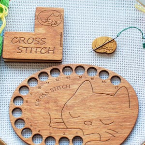 May include: A selection of cross-stitch tools and embroidery threads on white fabric. The wooden tools include a cat-shaped thread organizer, a needle minder, and a floss holder with the words "CROSS STITCH". Embroidery threads in yellow, green, and red.