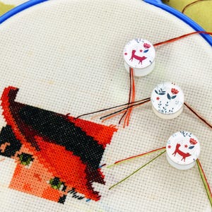 May include: Cross-stitch embroidery in a blue hoop. The design features a fox with red and black thread. Three white bobbins with floral and fox designs hold threads. The needlework is in progress, with various colored threads.