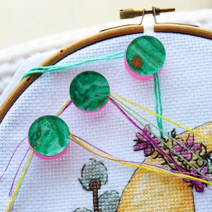 Peut inclure: Trois aimants ronds, verts et dorés, avec des bases roses, maintiennent des fils à broder sur un projet de point de croix. Le projet est tendu dans un cercle à broder en bois, avec des fils colorés visibles.