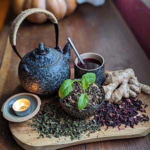 Pode incluir: Uma tábua de madeira com um bule preto, uma xícara de chá vermelho, uma tigela de ervas secas, um pedaço de gengibre e uma vela pequena.