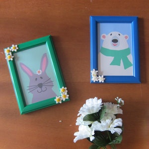 May include: Two picture frames with green and blue frames decorated with white flowers. The green frame has a picture of a grey rabbit with a pink nose and a yellow flower on its head. The blue frame has a picture of a white polar bear wearing a green scarf.