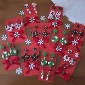 Peut inclure: Assortiment de boucles d'oreilles en argile polymère sur le thème de Noël présentées sur des cartes rouges avec des nœuds en velours rouge. Les boucles d'oreilles présentent des motifs tels que des étoiles, le Père Noël et des cannes en sucre.