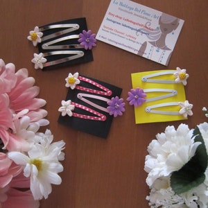 Puede incluir: Tres juegos de pinzas para el pelo con adornos de flores. Las pinzas son plateadas y tienen un patrón de lunares blancos, rosas y amarillos. Las flores son blancas con centros amarillos y moradas con centros amarillos.