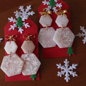 Könnte beinhalten: Ein Paar weiße Ohrringe mit Schneeflockenmuster, goldenen Schleifen und sechseckiger Form. Die Ohrringe werden auf roten Karten mit weißen Schneeflocken-Dekorationen präsentiert.