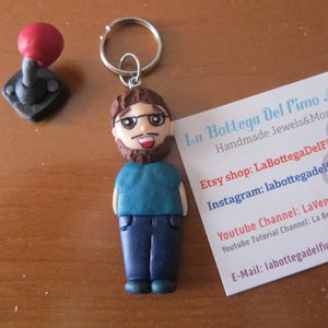 Pode incluir: Um chaveiro artesanal com uma figura em miniatura de um homem com óculos, barba e camisa azul, juntamente com um joystick vermelho. O chaveiro inclui um anel de metal e um cartão de visita com o texto "La Bottega Del Fimo Art".