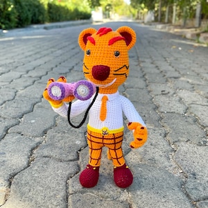 May include: A crocheted orange and white tiger plush toy wearing a white shirt with a red tie and holding a pair of purple binoculars.
