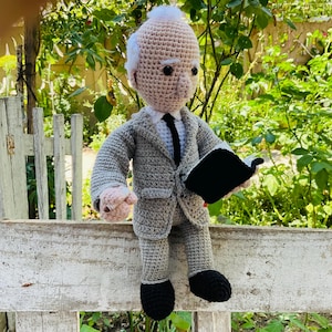 May include: A handmade crochet doll depicting a man in a grey suit, white shirt, and black tie, holding a black book. The doll has white hair and is seated on a white wooden surface. Green foliage forms the background.