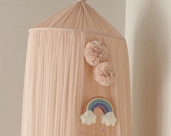 May include: A blush pink canopy with a white crib underneath. The canopy has a rainbow mobile hanging from it with white clouds and pink raindrops. There are two large pink pom poms on the canopy.