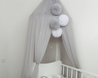 May include: A light gray tulle canopy with white and gray pom poms hangs over a white crib with a pink and gray braided bumper.