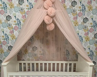May include: A white crib with a pink tulle canopy and pom-poms hanging from the top. The crib is against a floral wallpaper with the text "Powder Pink" written on the floor in front of the crib.