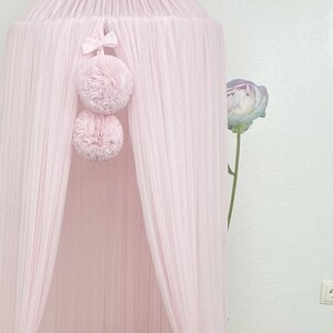 May include: A pink tulle canopy with two pink pom-poms hanging from the top. The canopy is draped over a white cot with a pink butterfly pillow inside.