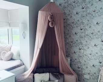 May include: A dusty pink canopy bed with a fluffy rug and pillows. The canopy is made of sheer fabric and has a pom-pom trim. The bed is set up in a corner of a room with a floral wallpaper.