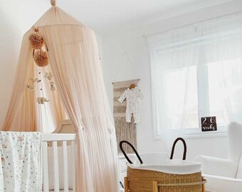 May include: A white cot with a pink canopy and a white and grey patterned blanket. A wicker Moses basket with a white liner sits on a wooden stand. A white rug is on the floor. The text "26 weeks" is visible on a sign in the background.