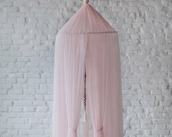 May include: A pink tulle canopy with a white tassel hanging from the center. The canopy is set up in a room with a white brick wall and a white wooden floor. There are white and brown pom-poms and a white cloud-shaped pillow on the floor.