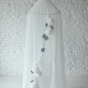May include: A white canopy bed with sheer fabric and a decorative garland of white pom-poms and blue stars. The canopy is suspended from the ceiling, creating a dreamy and whimsical atmosphere, perfect for a nursery or bedroom.