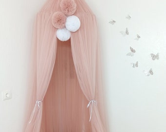 May include: A pink tulle canopy with pom-pom accents, a braided bumper, and a heart-shaped pillow. The canopy is suspended from the ceiling, creating a cozy space. A white faux fur rug sits beneath the canopy.